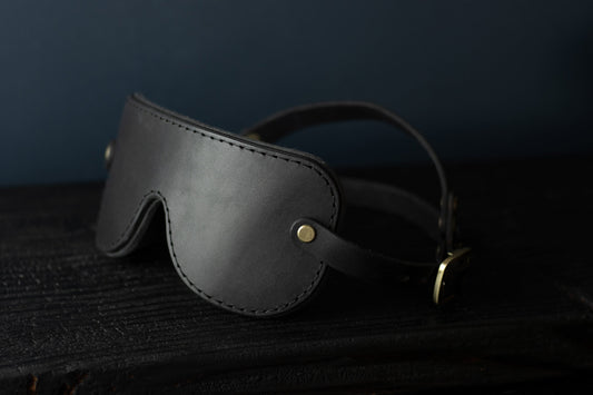 A black leather blindfold sits on a black surface. In the background, the double-strap fit for a secure hold during bdsm play is visible. There are bronze fittings and buckles on each side of the blindfold. 