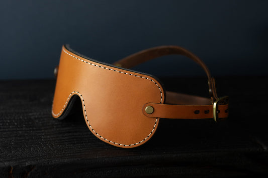 A cognac brown leather blindfold sits on a black surface. In the background, the double-strap fit for a secure hold during bdsm play is visible. There are bronze fittings and buckles on each side of the blindfold.