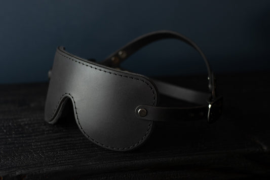 A black leather blindfold sits on a black surface. In the background, the double-strap fit for a secure hold during bdsm play is visible. There are gunmetal black fittings and buckles on each side of the blindfold. 