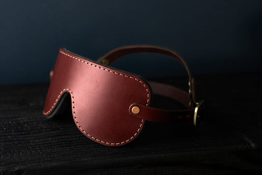 An oxblood red leather blindfold sits on a black surface. In the background, the double-strap fit for a secure hold during bdsm play is visible. There are bronze fittings and buckles on each side of the blindfold. 