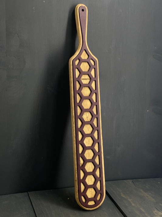 Image of the spanking paddle leaning against a black wall. The oak is visible through the honeycomb leather pattern overlay.