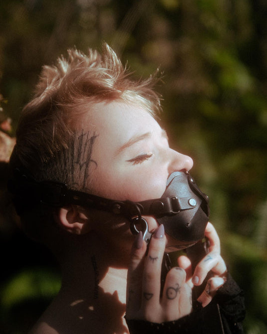 An individual models the muzzle gag, with a look of bliss on their face as they point their face towards sunlight. They are touching the muddle gag gently with their fingertips. The gag covers from underneath their nose, cupping their chin gently.