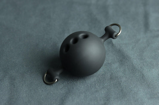 A black silicone ballgag sits on a grey piece of fabric. On each side are D rings for clipping it into a leather ball gag harness. 