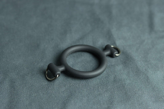 A ring gag sits on a grey piece of fabric. On each side, there is a D ring for locking into a leather ballgag head harness.