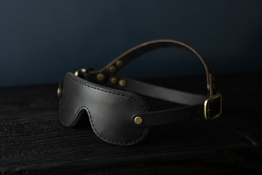 A petite black leather blindfold with bronze hardware sits on a black wood surface. The double strapped head harness is visible.