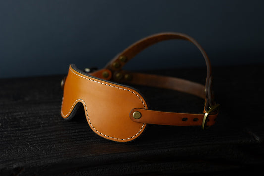 A petite cognac brown leather blindfold with bronze hardware sits on a black wood surface. The double strapped head harness is visible. 