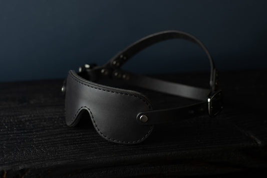 A petite black leather blindfold with black hardware sits on a black wood surface. The double strapped head harness is visible. 