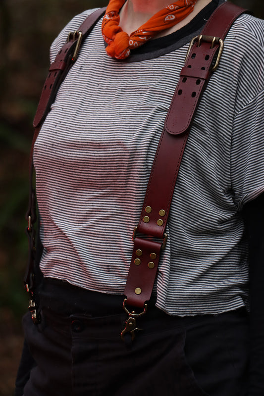 An individual is wearing the leather suspenders over a striped black tee shirt, with black pants and an orange scarf. The suspenders feature a beautiful and functional buckle that rests near the shoulders, brass clips at the bottom, and brass rivets.