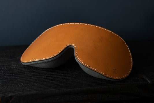 A cognac brown leather blindfold with a weighted flaxseed back sits on a black piece of wood.  The stitching is tan.